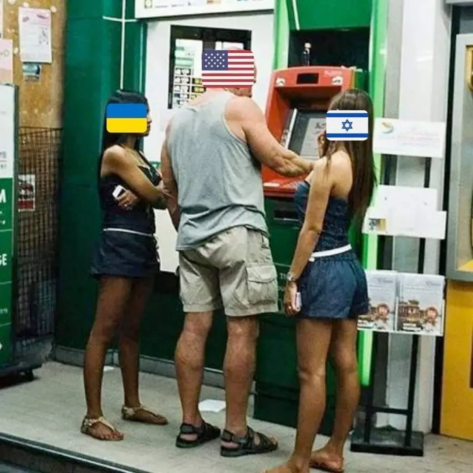 Three people stand at an atm; the man in the center wears a gray tank top and khaki shorts, with an american flag over his…