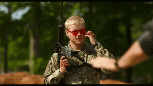A soldier in camouflage and red-tinted glasses holds a rifle while another person's arm is visible. The soldier initially…