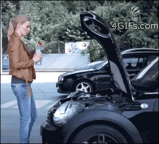 A woman in a brown jacket and jeans stands beside a black car with its hood open. She holds a water bottle and proceeds to…