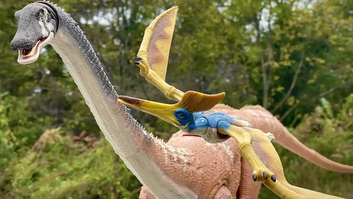 A toy pterosaur with blue and yellow coloring is perched on the neck of a larger toy dinosaur, which has a long neck and…