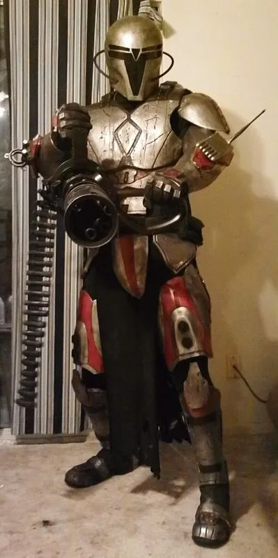 A person in a full Mandalorian armor costume, resembling the character from Star Wars, stands indoors holding a large…