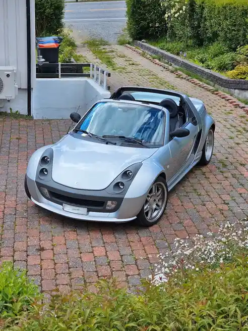 A silver Smart Roadster convertible is parked on a red brick driveway surrounded by greenery and a white house. The car has…