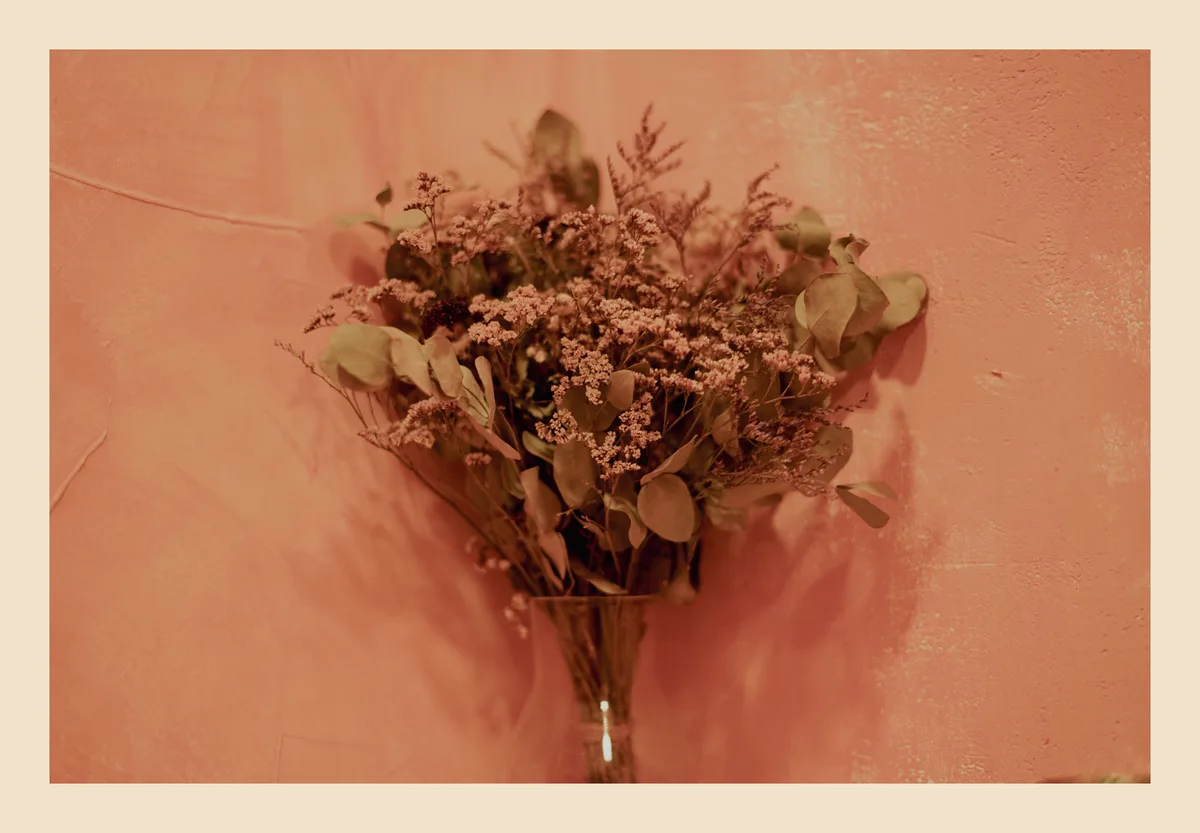 A still life photo shows a bouquet of dried flowers and eucalyptus leaves in a glass vase, set against a textured…