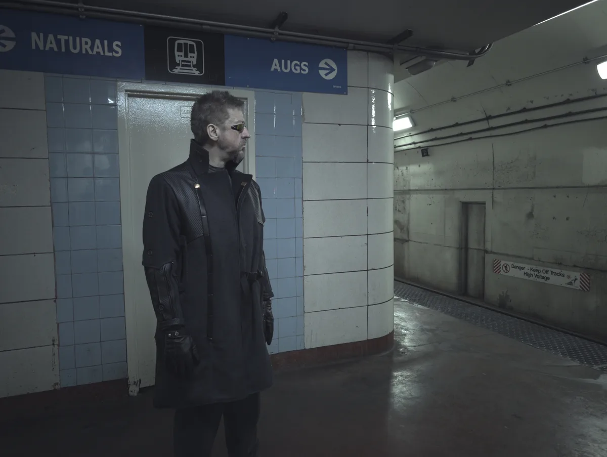 A man in a dark trench coat and sunglasses stands in a dimly lit subway station. Above him, blue signs read 'NATURALS' and…
