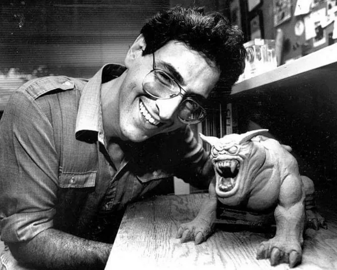 A black-and-white photo shows a smiling man with glasses leaning over a table, next to a detailed, roaring goblin figurine…