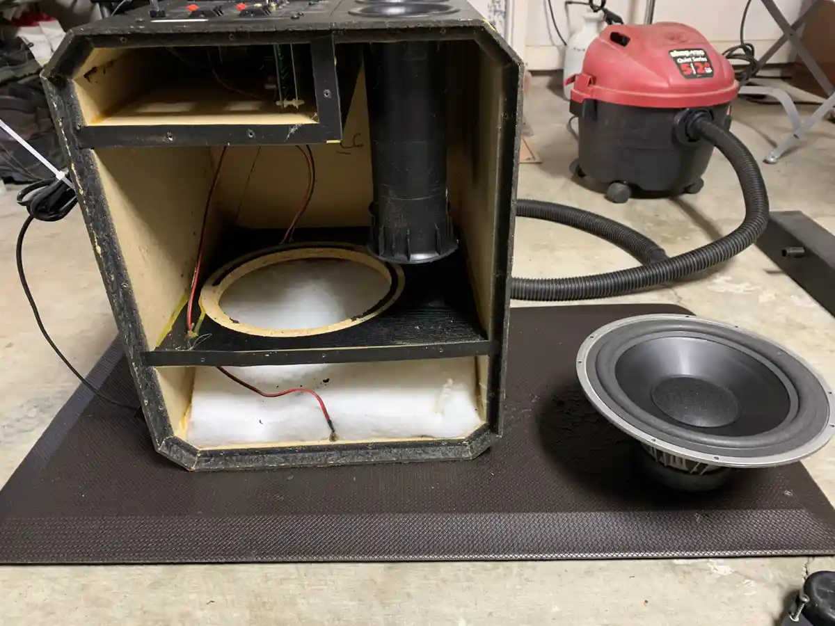 A partially assembled subwoofer enclosure with exposed wiring and a circular cutout sits next to a large subwoofer driver on…