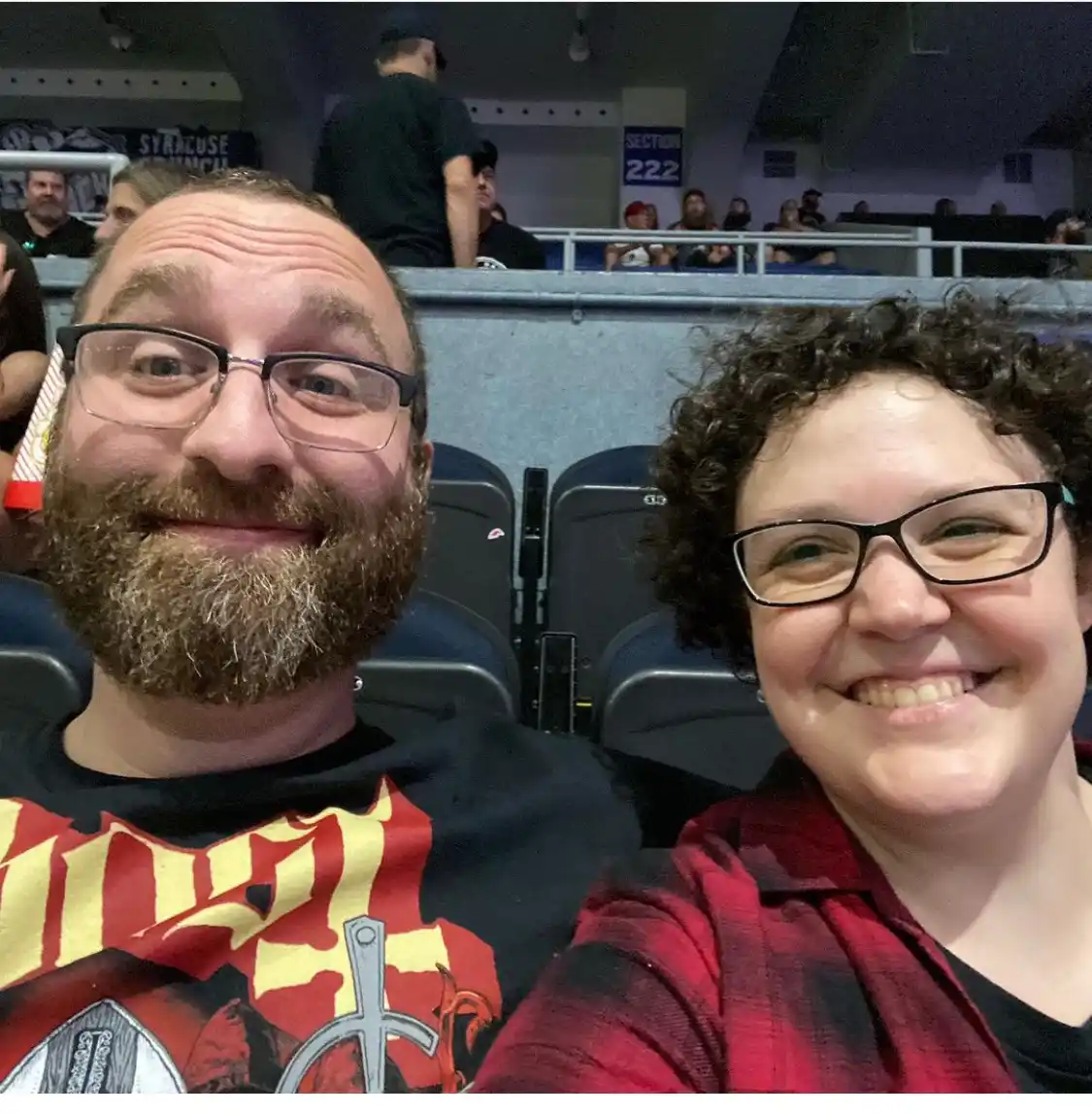A selfie of a smiling man with a beard and glasses and a smiling woman with curly hair and glasses, taken in stadium…