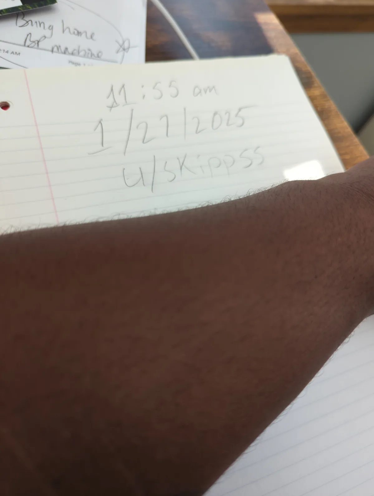 A close-up photo shows a person's arm resting over a lined notebook. The notebook has handwritten text in pencil that reads…