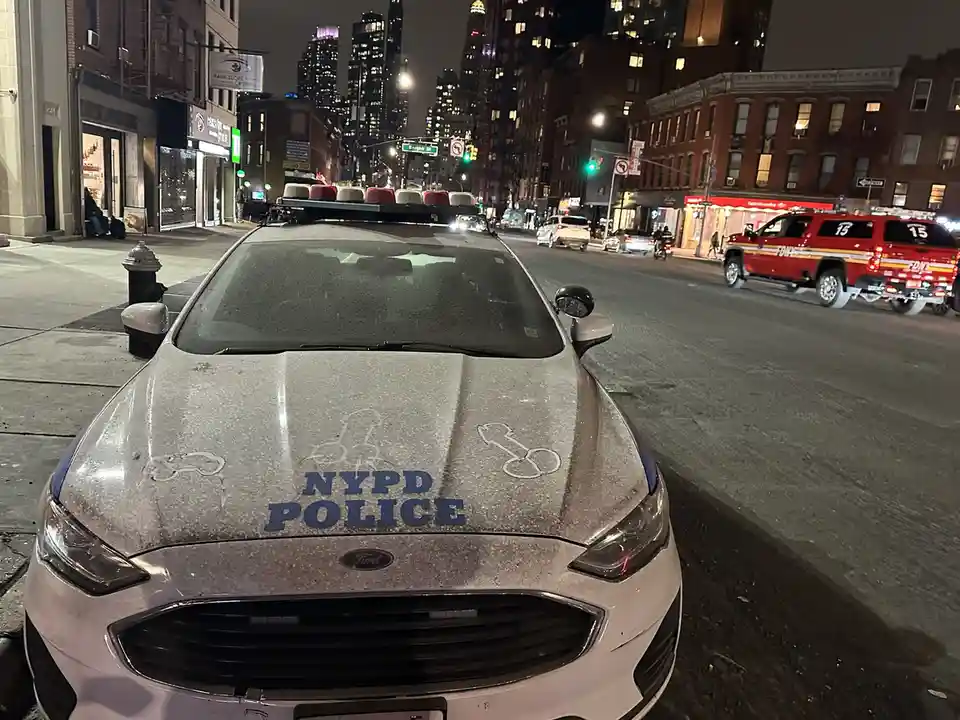 A front-facing photo of an NYPD police car parked on a city street at night, covered in a layer of snow or frost. The car's…