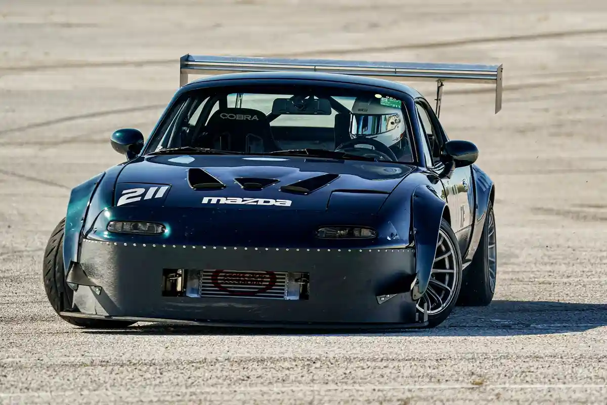 A dark blue Mazda race car with the number 211 on the hood, a large rear wing, and a driver in a helmet, is shown on a…