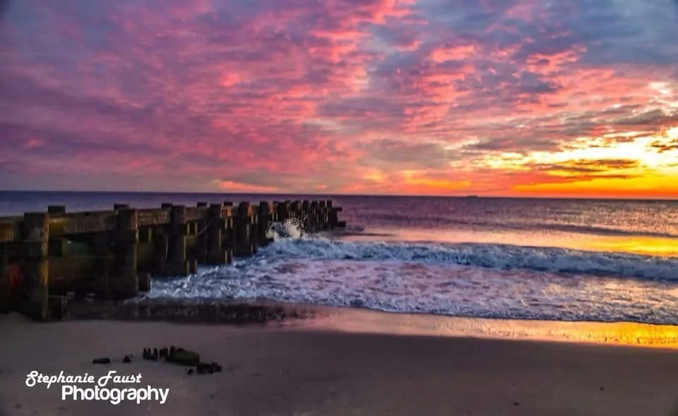 A vibrant sunset over a beach with waves crashing against a wooden pier, the sky painted in pink, orange, and purple hues…