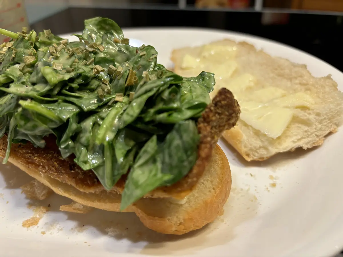 A close-up photo shows a sandwich on a white plate, with one half topped with wilted spinach and chopped green herbs, and…