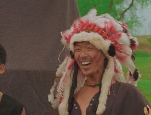 A man with a wide, joyful laugh wears a large, elaborate headdress with red and white feathers and fur trim. He has a green…