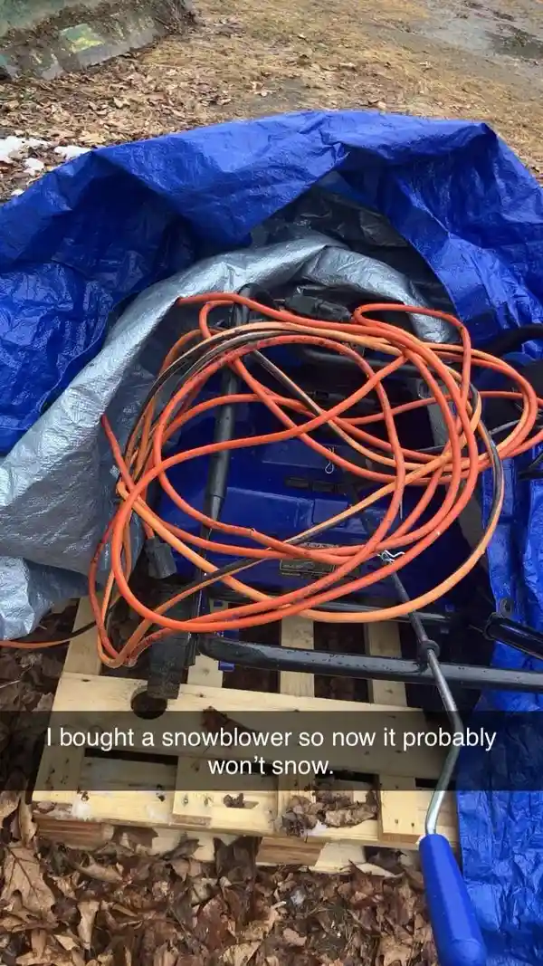 A blue snowblower is covered with a blue tarp and a silver tarp, sitting on a wooden pallet surrounded by dry leaves and a…