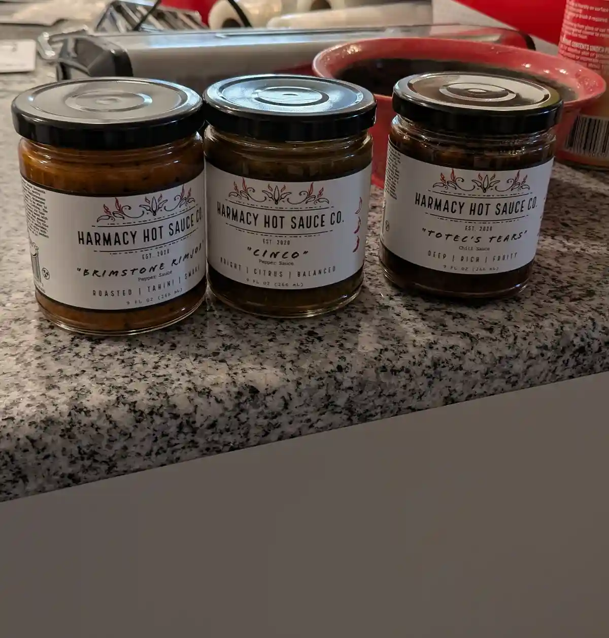Three jars of Harmacy Hot Sauce Co. sit on a granite countertop, each labeled with a unique name: 'Brimstone Kimjob'…