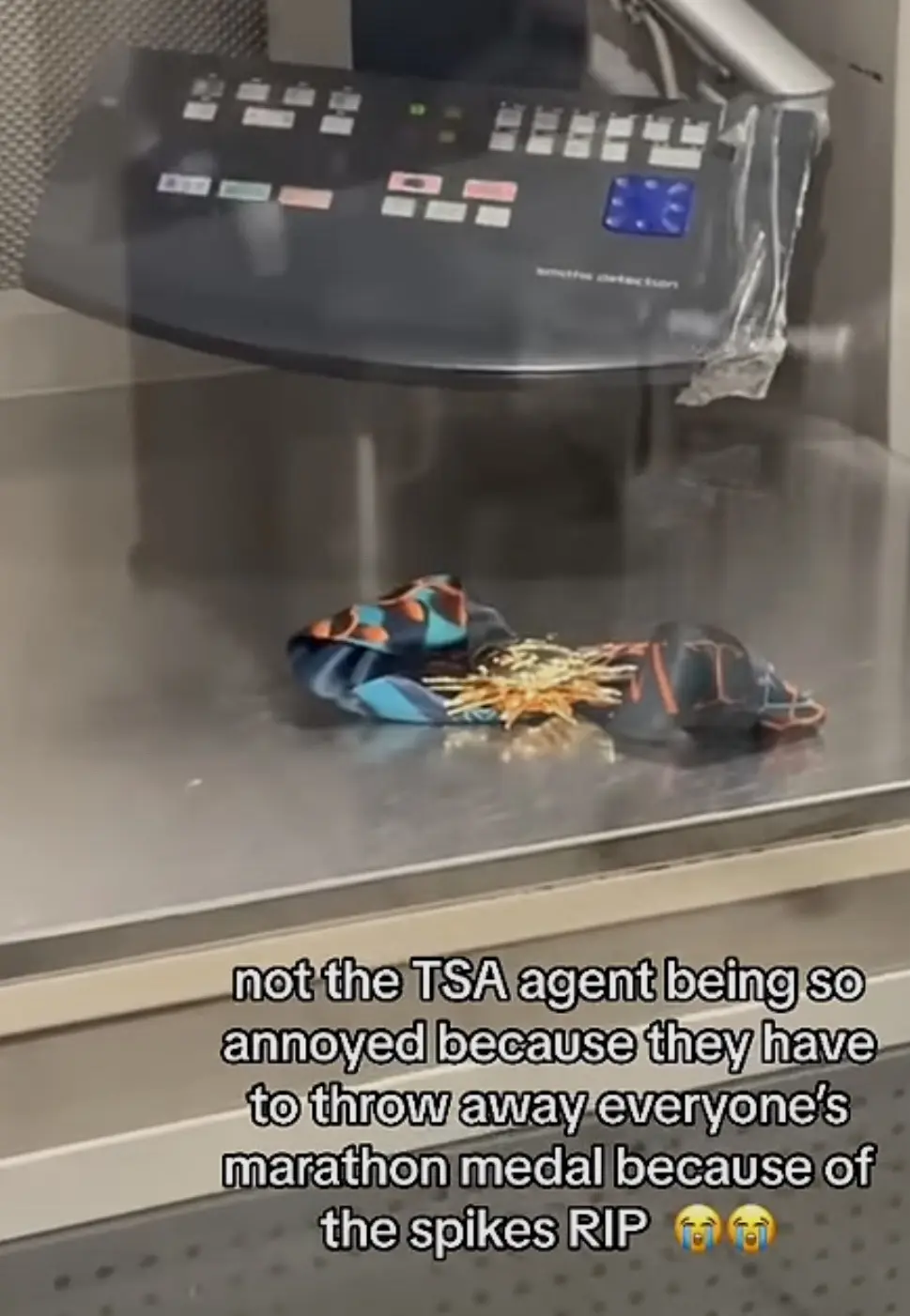 A close-up shot of a metal detector tray at an airport security checkpoint, where a crumpled marathon medal with spikes is…