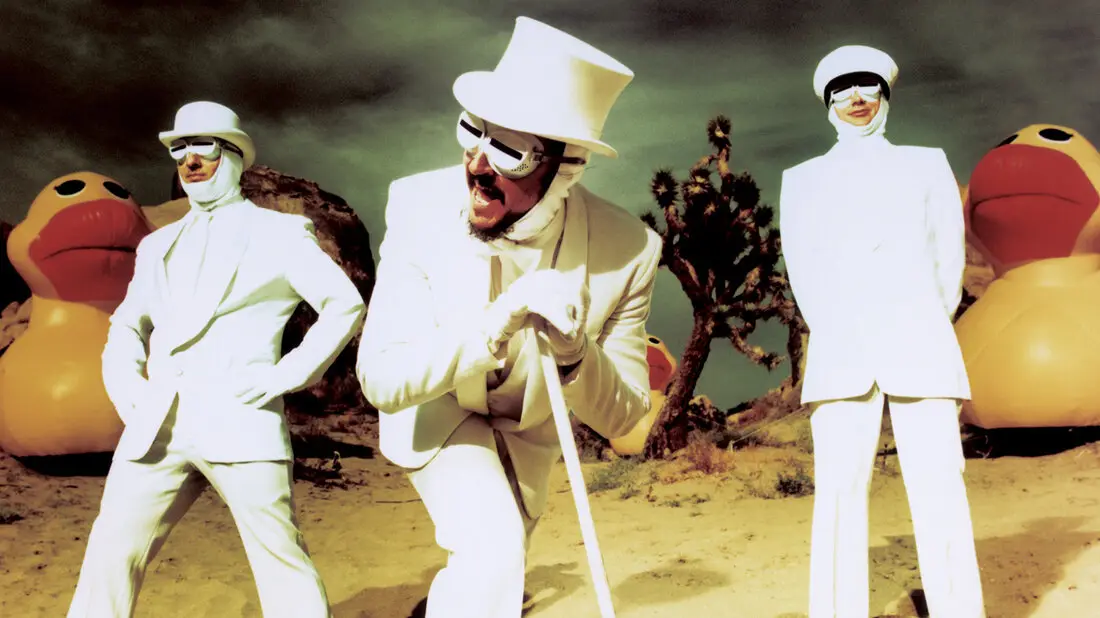 three men in white suits, white hats, and white face coverings stand in a desert with giant rubber ducks beside them. The…