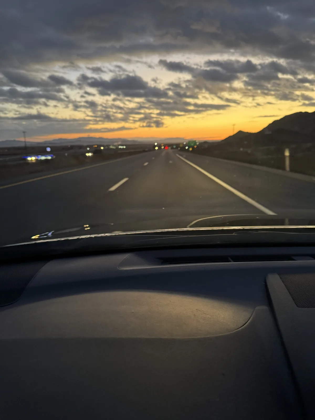 A photo taken from inside a car looking out at a highway during sunset or sunrise, with orange and gray clouds above and…