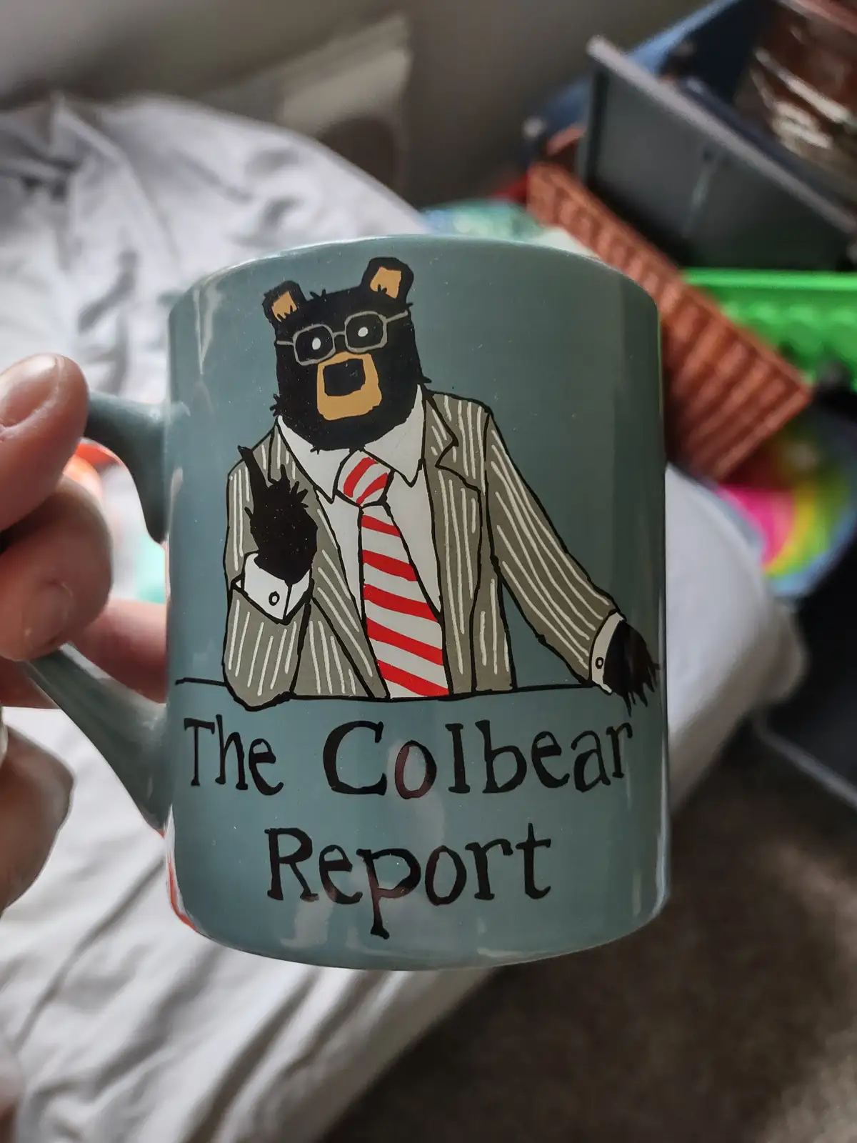 A person holds a light blue mug featuring a cartoon black bear wearing glasses, a pinstripe suit, and a red-and-white…