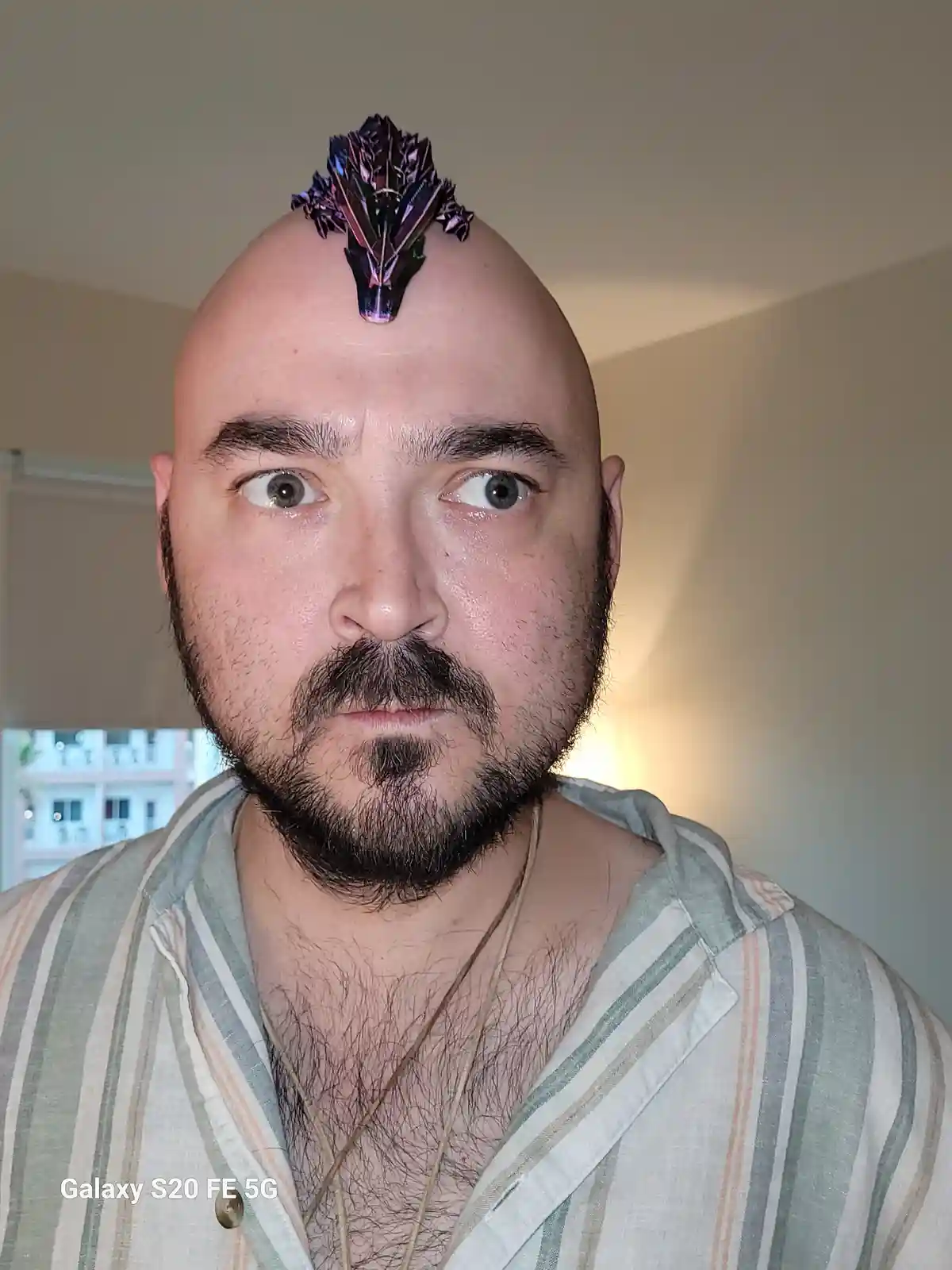 A bald man with a goatee and wide-eyed expression stares at the camera, with a shiny, iridescent, spiky purple and blue…
