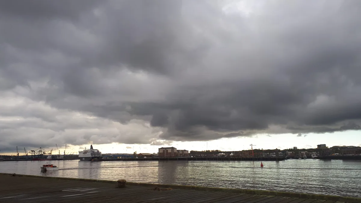 A wide shot of a harbor under a heavy, dark, and dramatic cloud cover, with a large ferry docked and smaller boats on the…