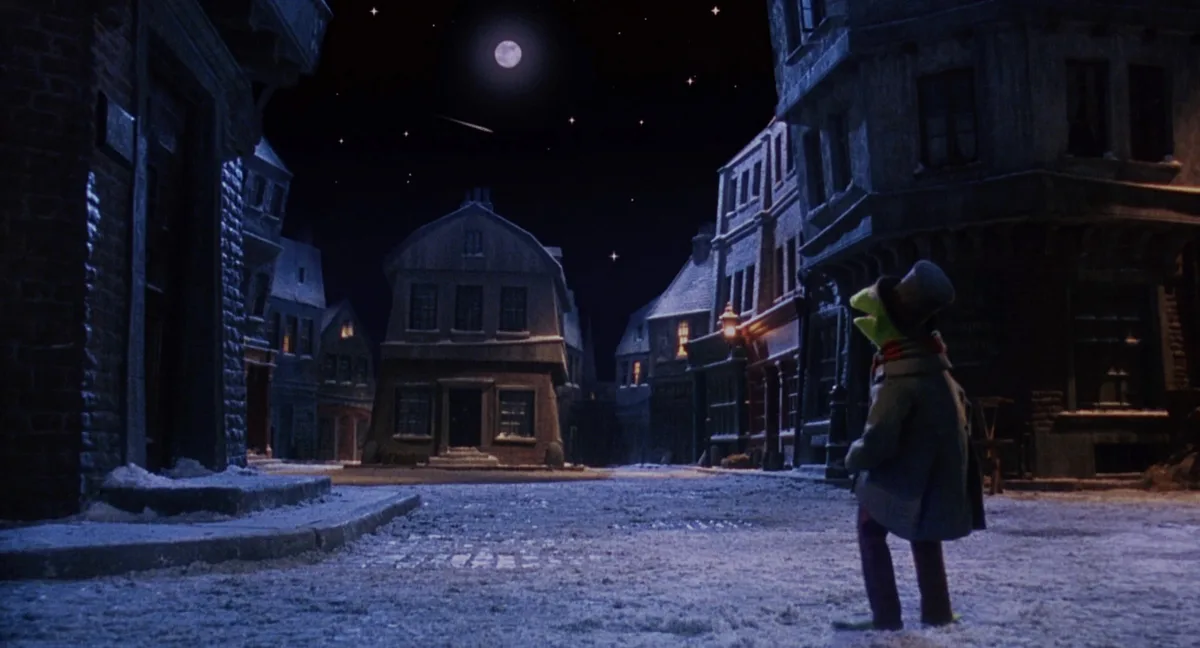 A still from 'The Muppet Christmas Carol' shows Kermit the Frog standing alone in a snowy, moonlit street at night, wearing…