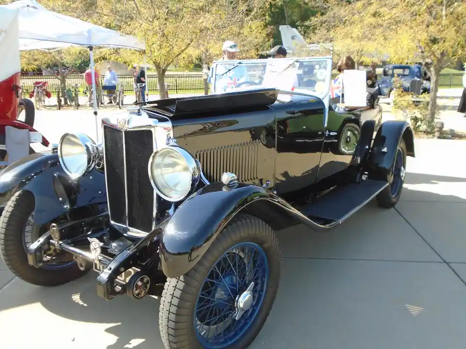 A shiny black vintage MG convertible with blue wire wheels is parked at an outdoor car show, with people and other classic…