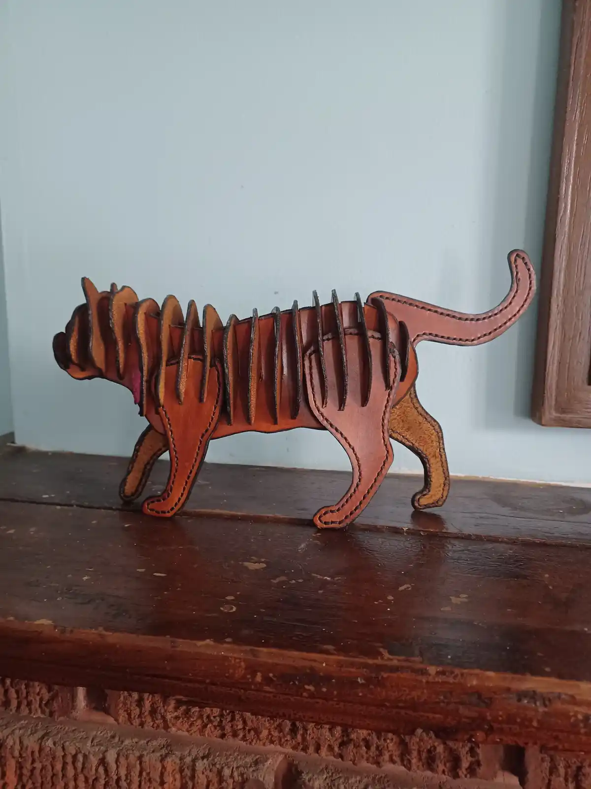 A handmade leather cat figurine with its spine visibly segmented into individual, slightly raised slats, sitting on a wooden…