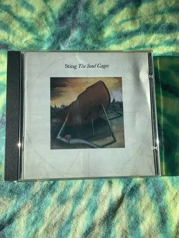A scratched CD case for Sting's album 'The Soul Cages' rests on a tie-dye fabric. The cover art depicts a large, brown…