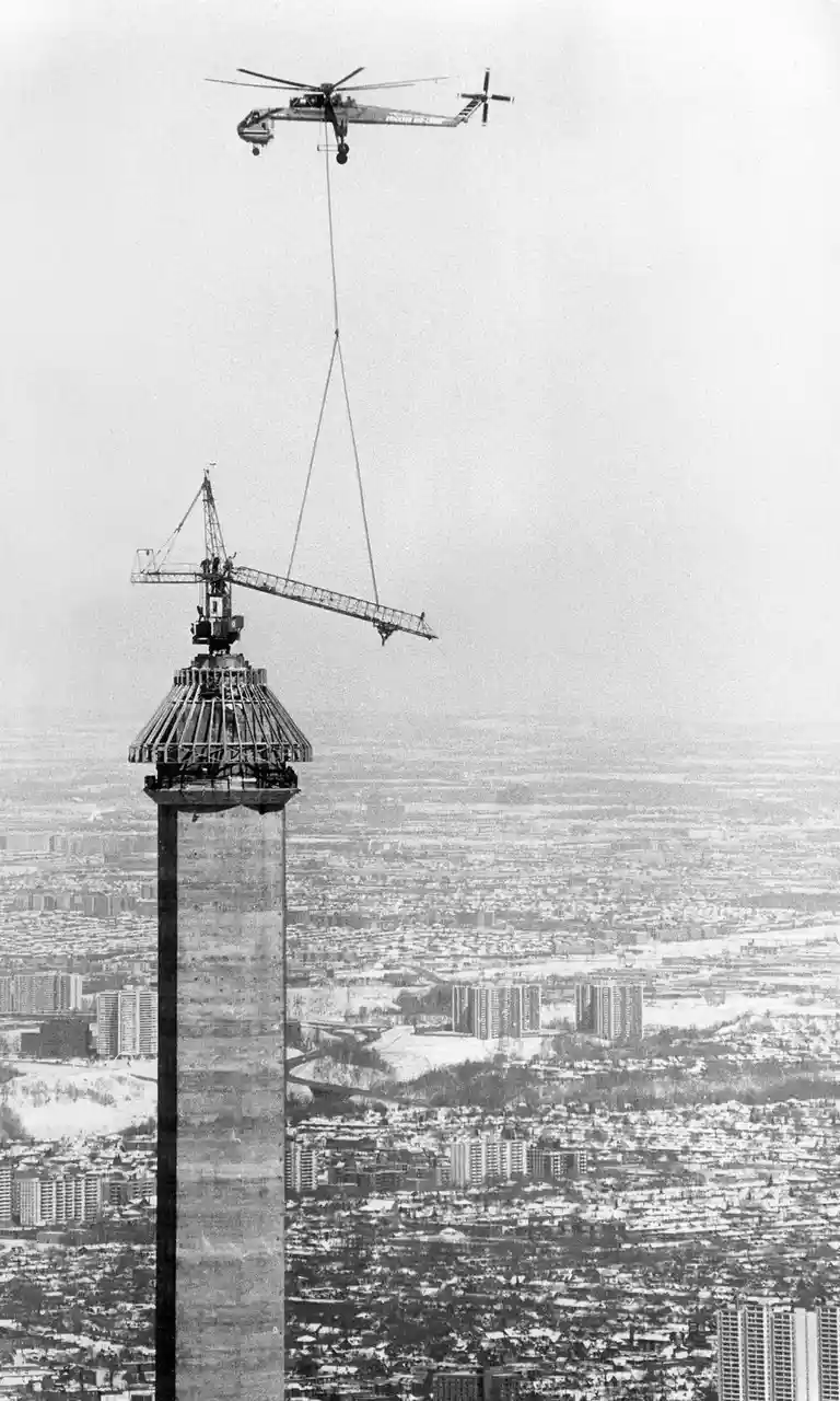 A black-and-white photo shows a helicopter suspended above a tall tower, using cables to lift a large crane that is being…