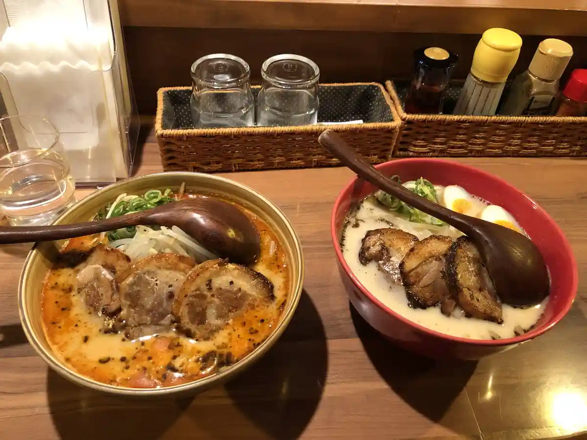 Two bowls of ramen sit side by side on a wooden table, one with a reddish broth and the other with a creamy white broth…