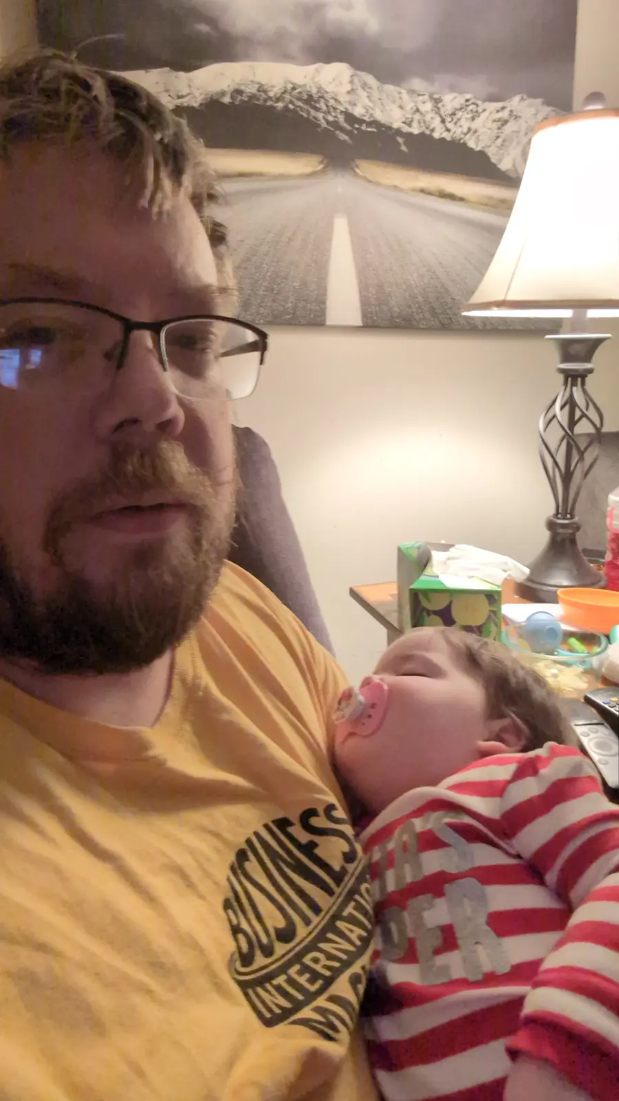 A man with glasses and a beard wearing a yellow t-shirt holds a sleeping baby in a red and white striped shirt with a…