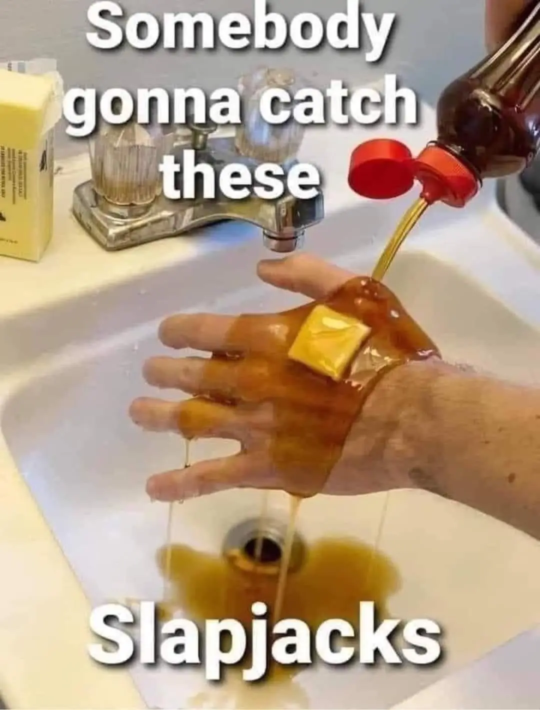 A hand is held under a sink faucet while syrup is poured from a bottle onto it, with a pat of butter resting on the hand…