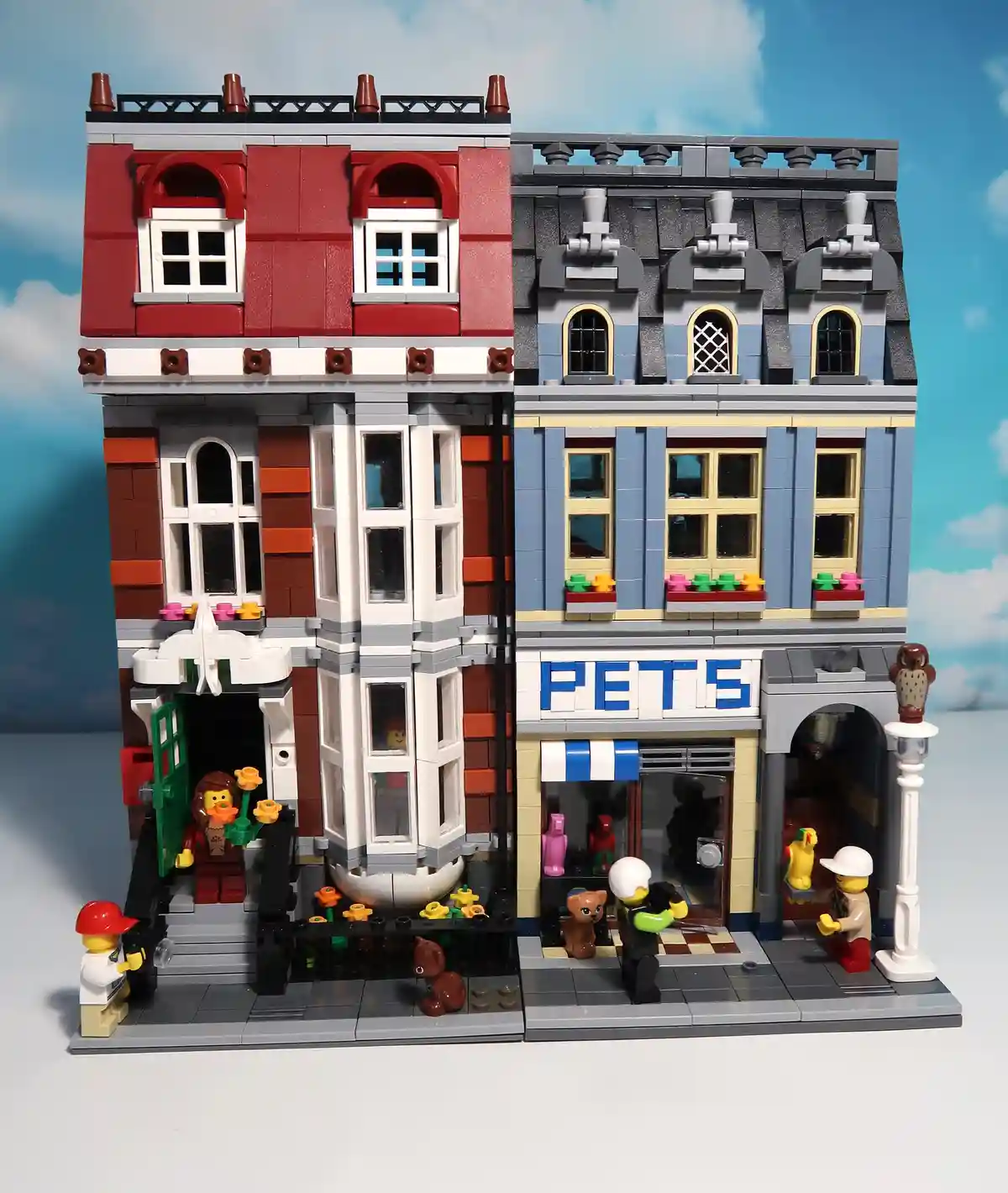 A detailed LEGO diorama shows two adjacent buildings: a red-brick house with a woman holding flowers at the entrance and a…