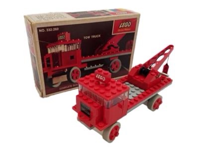 A red LEGO tow truck built from bricks sits in front of its original box, which shows an image of the same truck. The box is…