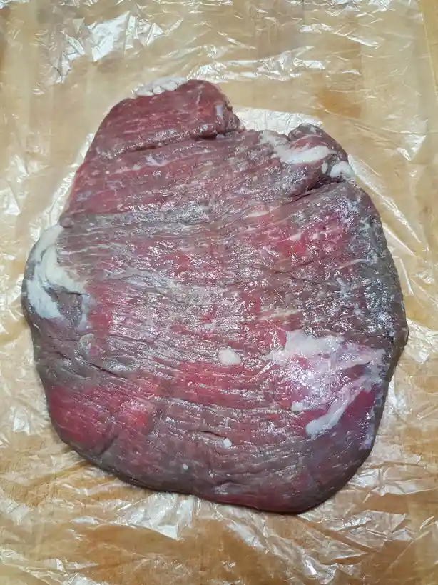 A large, raw, irregularly shaped cut of red meat with visible fat marbling rests on crinkled plastic wrap atop a wooden…