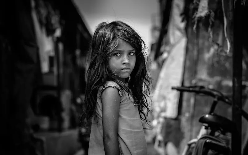 A black-and-white photograph of a young girl with long, messy hair standing in a narrow alleyway, looking directly at the…