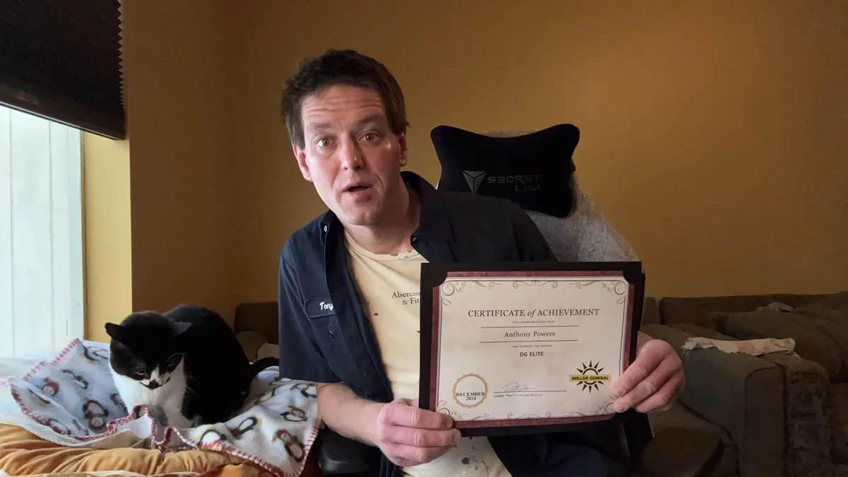 A man sits in a gaming chair holding a framed 'Certificate of Achievement' from Dollar General, which awards 'DG Elite'…