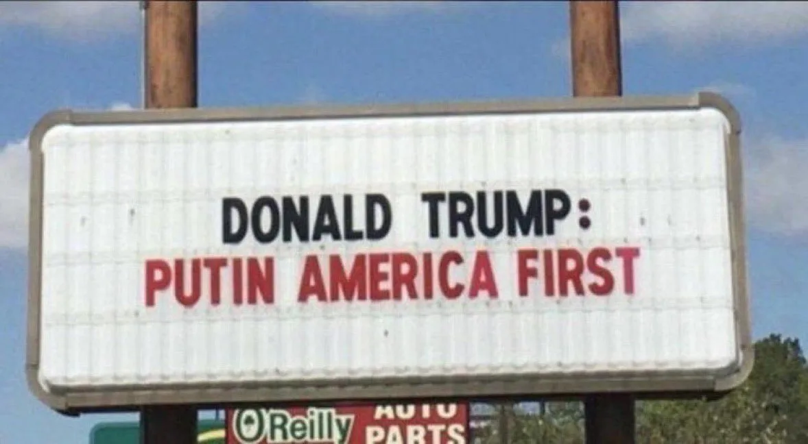 A white, corrugated metal sign with black and red lettering reads 'DONALD TRUMP: PUTIN AMERICA FIRST'. The sign is mounted…
