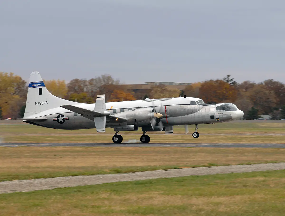 A silver twin-engine propeller plane, likely a Lockheed P-3 Orion, is shown taxiing on a runway with autumn trees in the…