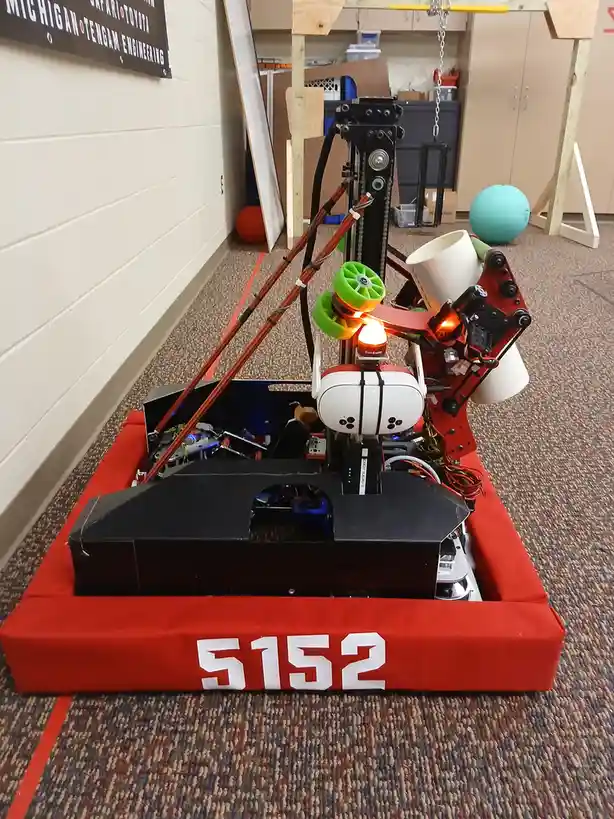 A complex, custom-built robot with exposed wiring, green wheels, and a white sensor module sits on a red padded base marked…