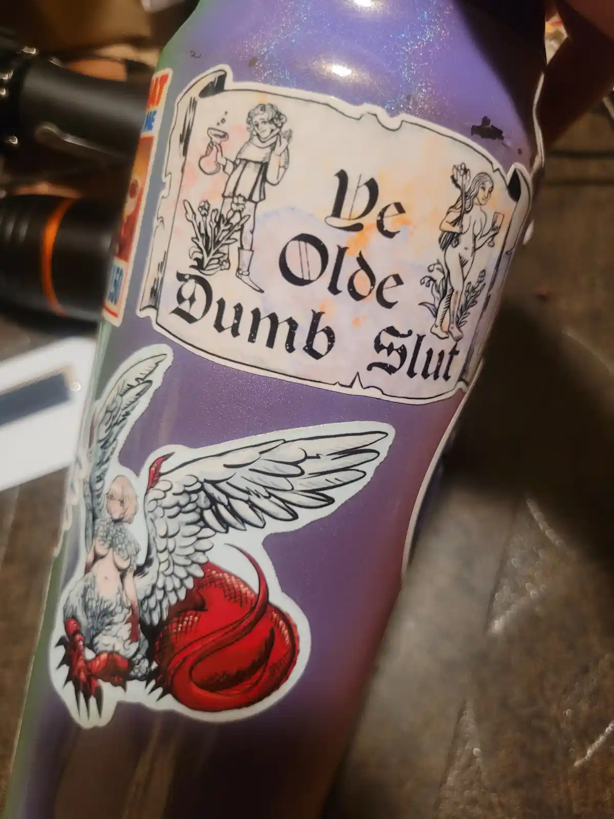 A close-up of a purple glittery bottle with a vintage-style label reading 'Ve Olde Dumb Slut' in gothic font, flanked by…