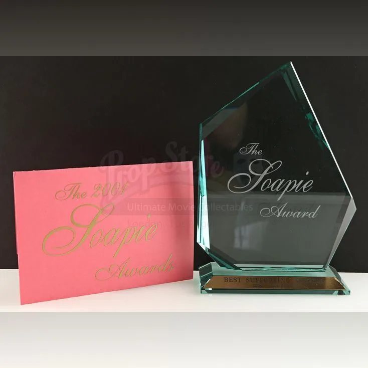 A glass award trophy and a pink envelope both engraved with 'The 2001 Soapie Awards' sit side by side on a white surface…