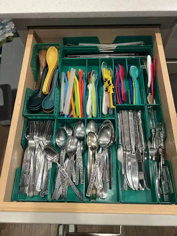 An overhead view of a kitchen drawer filled with green plastic organizers holding an assortment of cutlery, including adult…