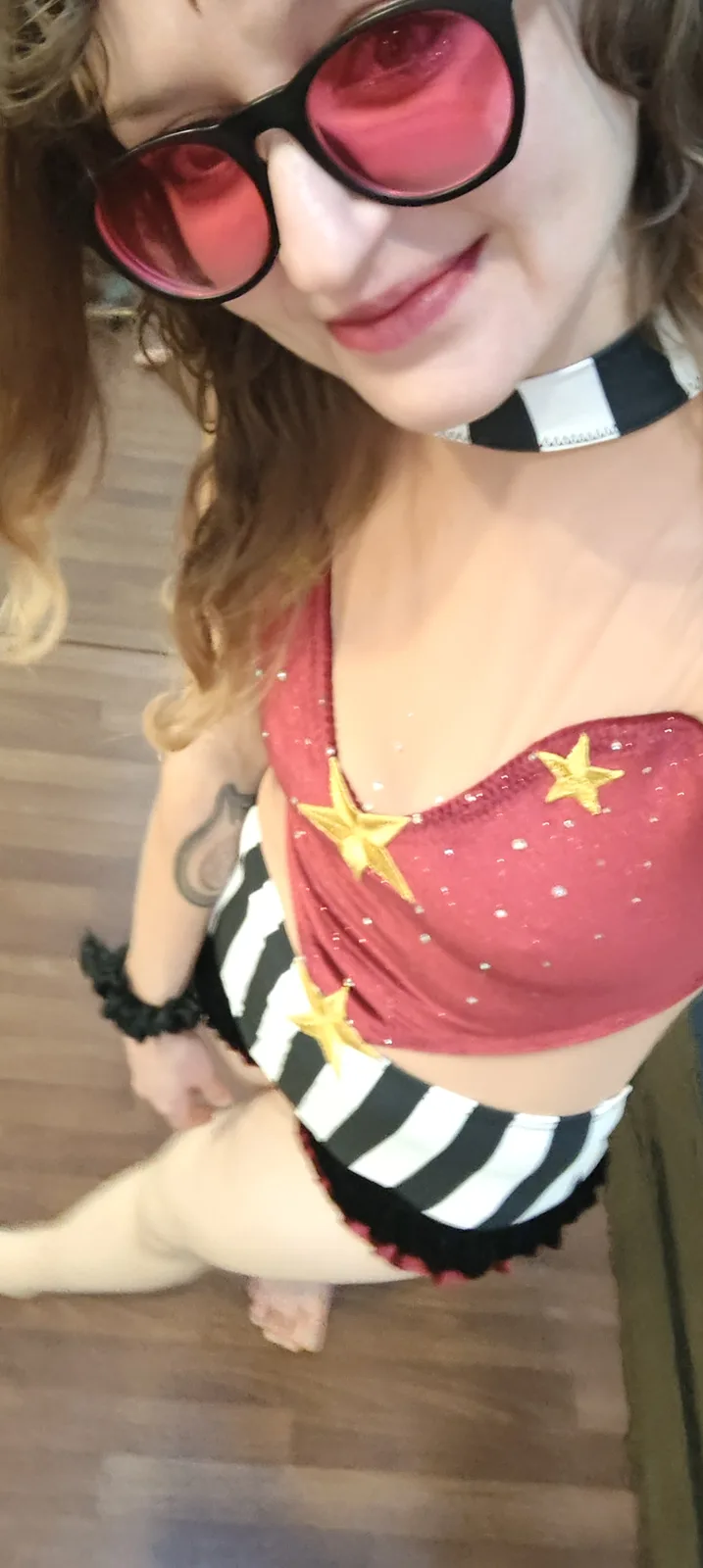 A person with curly hair wearing oversized pink-tinted sunglasses, a red sequined crop top with gold stars, and…