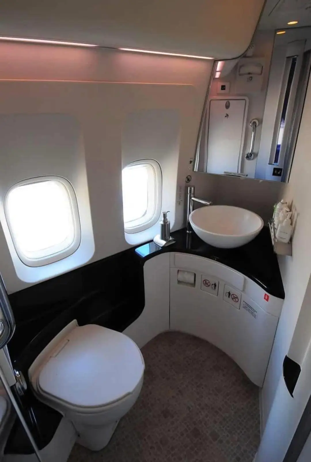 The image shows a compact, high-end airplane lavatory with two small windows, a white toilet, a modern bowl-shaped sink on a…