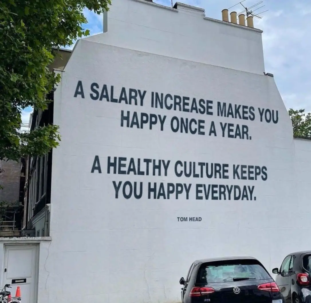 A large white building wall features bold black text stating 'A SALARY INCREASE MAKES YOU HAPPY ONCE A YEAR. A HEALTHY…