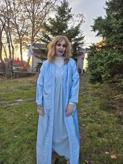 A person stands outdoors in a backyard wearing a light blue, quilted robe over a matching nightgown, with zombie makeup on…