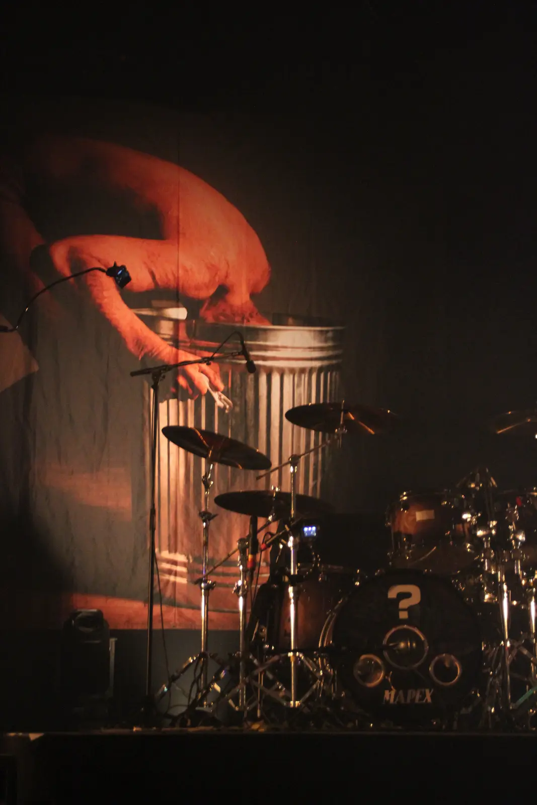 A dimly lit stage shows a drum kit in the foreground, with a large projection behind it of a person's arm reaching into a…