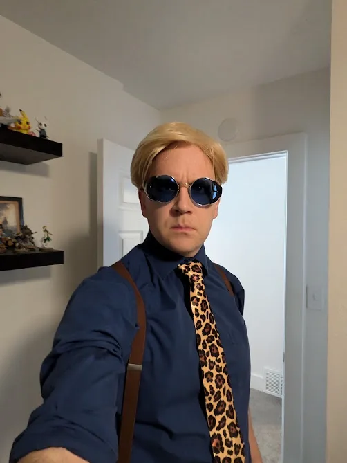 A person with blonde hair, wearing round dark sunglasses, a blue button-up shirt, brown suspenders, and a leopard-print tie…